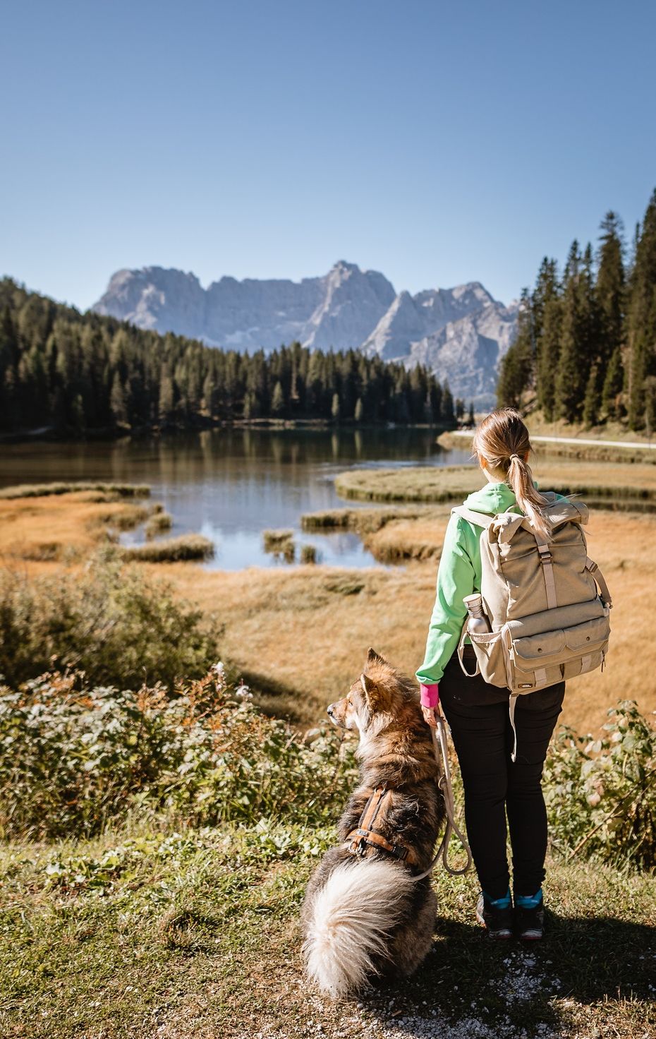Frau mit Rucksack und Hund vor einem See im Hintergrund Gebirge