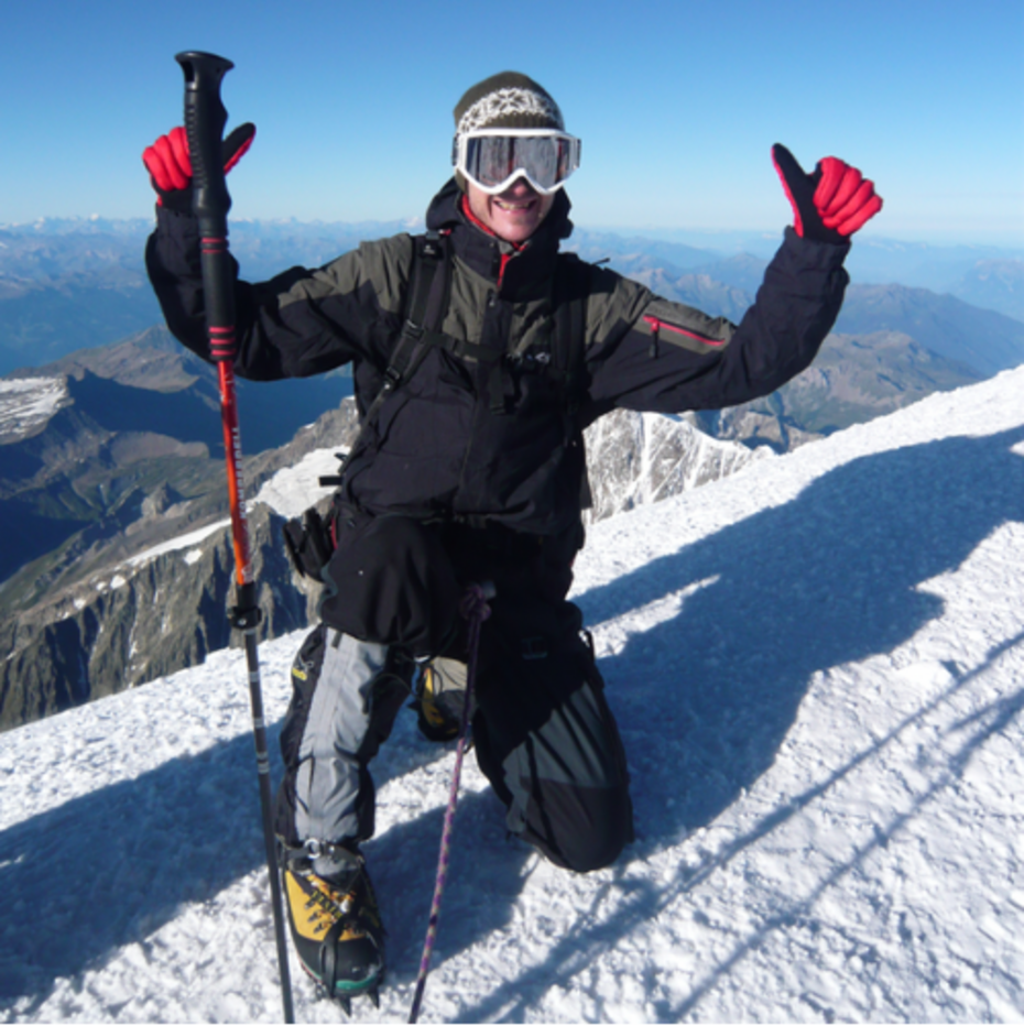 Ein Mann in hochalpiner Kleidung mit Steigeisen kniet im Schnee. Im Hintergrund eine Berglandschaft.