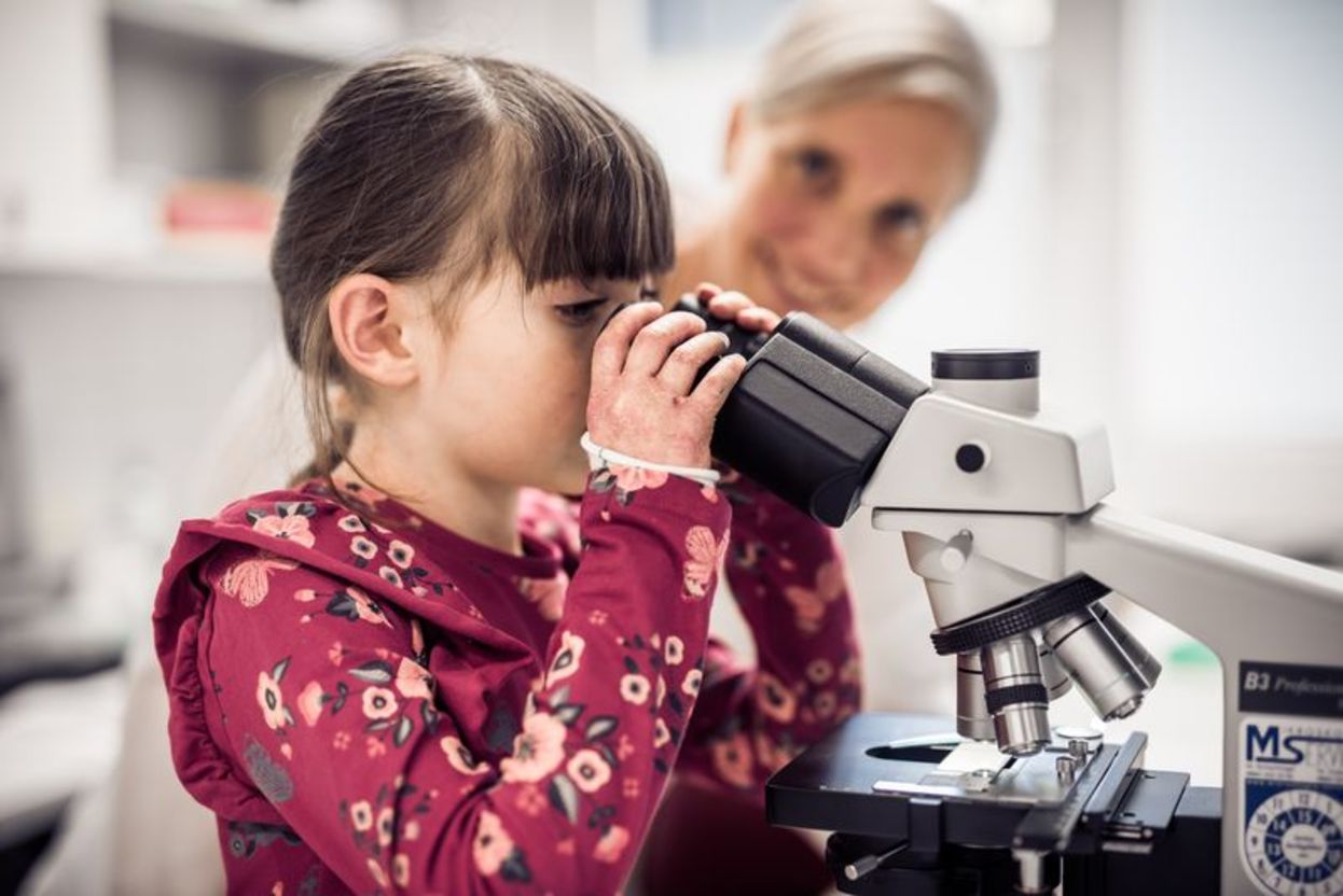 Schmetterlingskind schaut durch ein Mikroskop, im Hintergrund EB-Forscherin