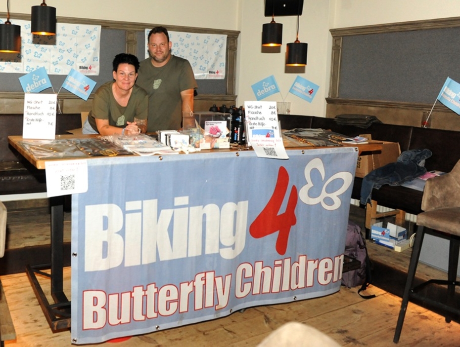 Ein Mann und eine Frau stehen hinter einem Tisch mit T-Shirts und einem Banner mit der Aufschrift Biking 4 butterlfy Children