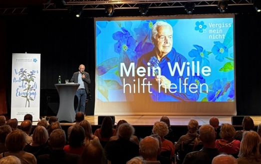 Projektion auf einer Leinwand, davor Menschen auf einer Bühne und Menschen im Publikum