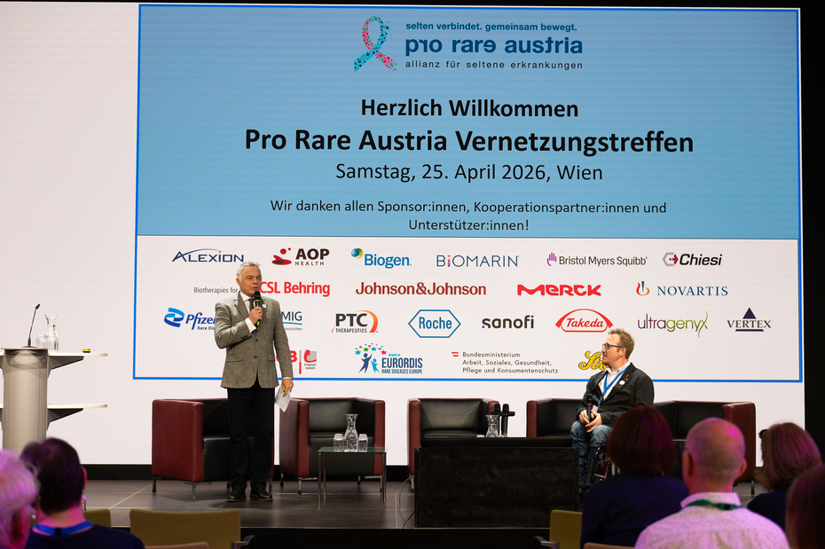 Ein Mann steht auf der Bühne vor einer großen Leinwand, im Vordergrund sieht man Publikum