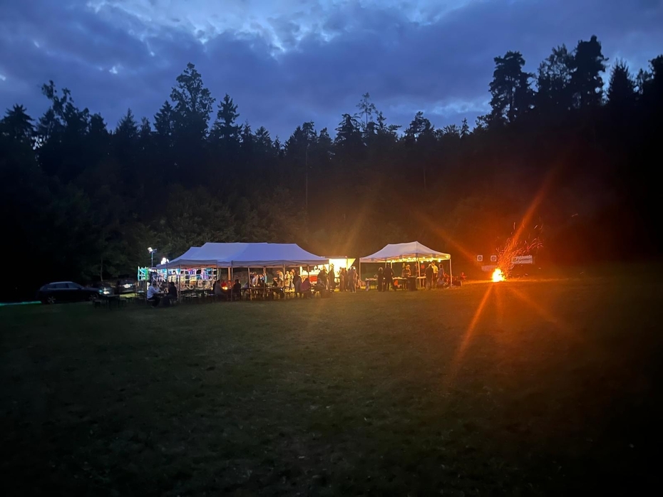 Zwei große Festzelte auf einer Wiese mit Wald im Hintergrund und Abendstimmung. Rechts im Bild ein großes Lagerfeuer.