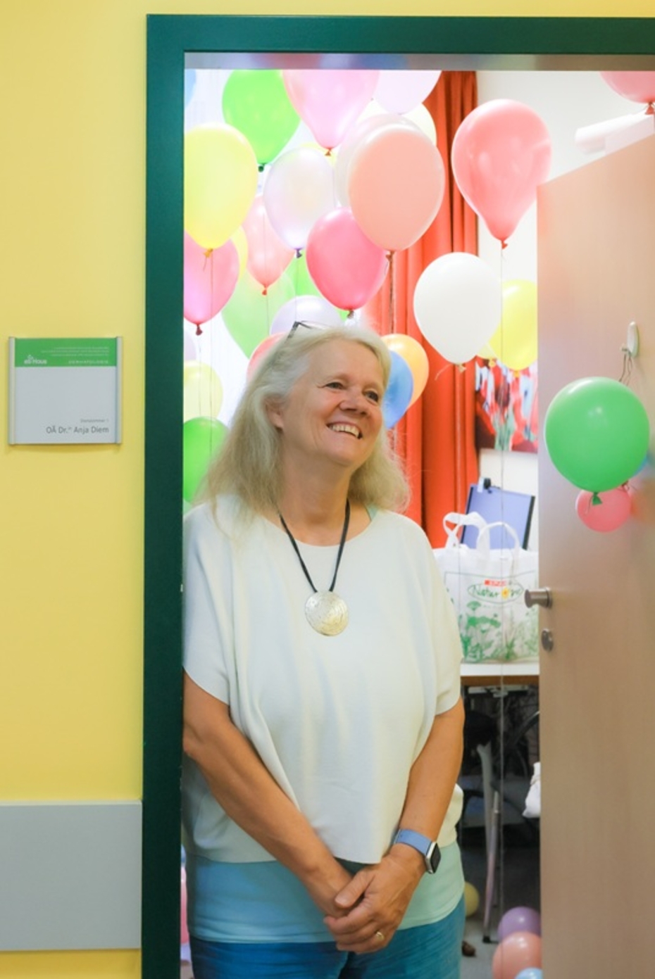 Frau mit weißem Kittel steht in einem Türrahmen, im Hintergrund viele bunte Luftballons