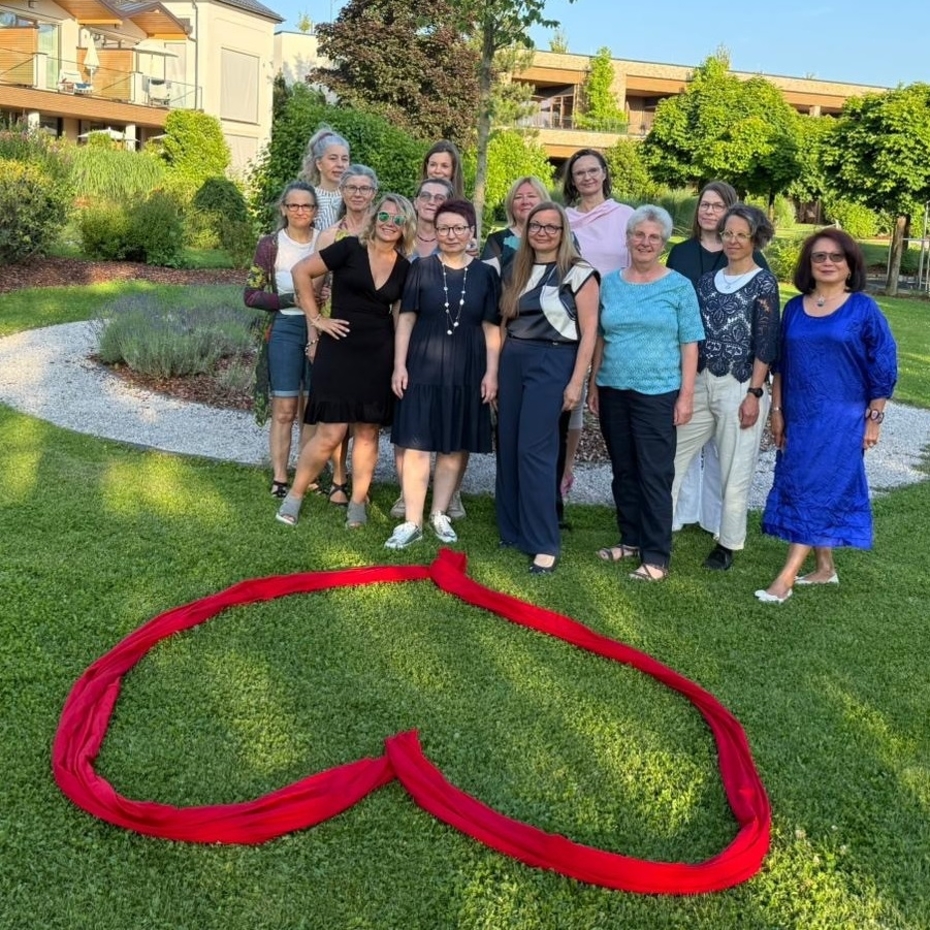 Gruppe von Frauen steht auf einer Wiese, vor ihnen liegt ein rotes Herz, aus einem Stoffband geformt