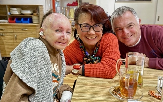 ein Schmetterlingskind im Rollstuhl, eine Frau und ein Mann sitzen an einem Tisch