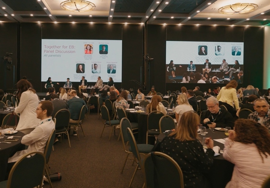 Menschen sitzen in einer Cafeteria, im Hintergrund werden Panels und Diskussionen auf Leinwänden übertragen