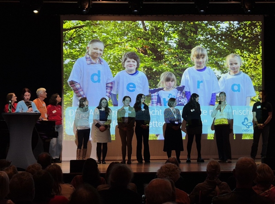 Menschen sitzen im Auditorium, auf der Bühne stehen weitere Menschen vor einer großen Leinwand, auf der Schmetterlingskinder abgebildet sind