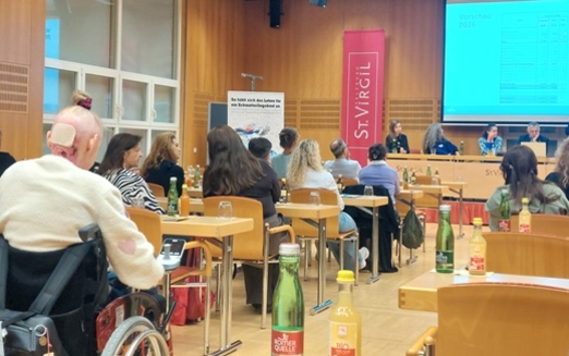 Menschen sitzen in einem Seminarraum, vorne ist eine Bühne und eine Leinwand